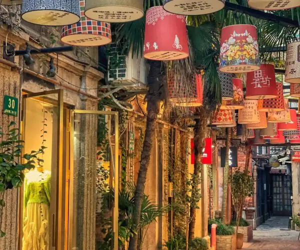 Narrow cobblestone street lined with boutique storefronts and potted plants, colorful lanterns hanging overhead.