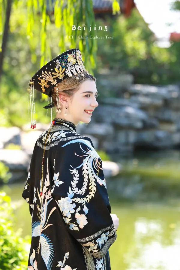 hanfu photoshoot, zhongshan park, beijing, female, solo, chinatrip, Woman in an ornate black headdress and traditional embroidered robe, smiling in a garden setting by a pond.