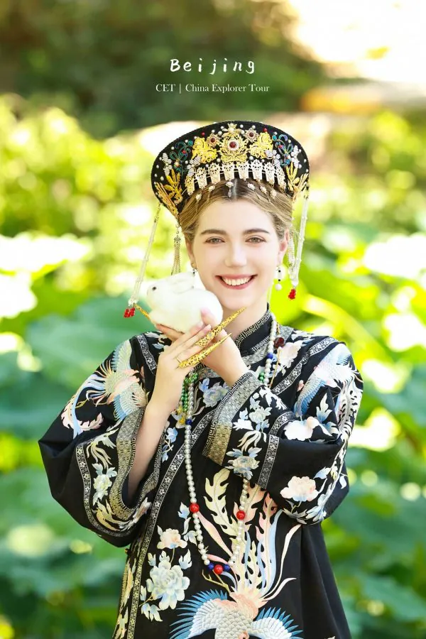 hanfu photoshoot, zhongshan park, beijing female, solo, chinatrip, Young woman in an ornate Chinese ceremonial headdress and embroidered robe, smiling while holding a white rabbit outdoors in a garden setting.