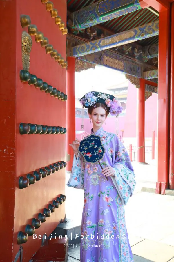 hanfu photoshoot, the forbidden city, beijing, qing dynasty, female, solo, chinatravel, Woman in a lavender traditional Chinese dress with a floral headdress posing beside a red ornate gate.