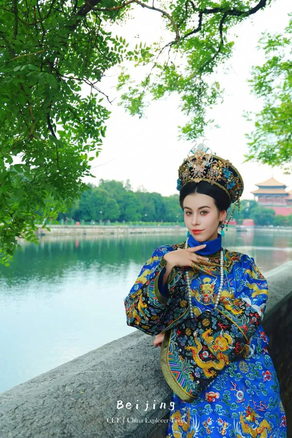 hanfu photoshoot, the forbidden city, beijing, qing dynasty, female, solo, chinatravel, Woman in an ornate embroidered Chinese robe and headdress posing beside a lake by green trees.