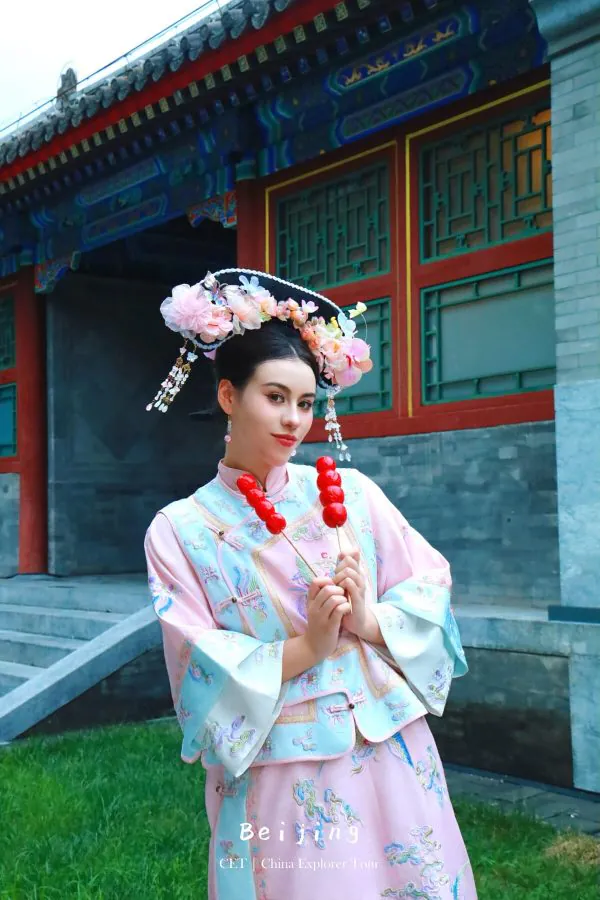 hanfu photoshoot, the forbidden city, beijing, qing dynasty, female, solo, chinatravel, Woman in pastel hanfu with a large flower headdress posing with red candy skewers in front of a traditional Chinese building.