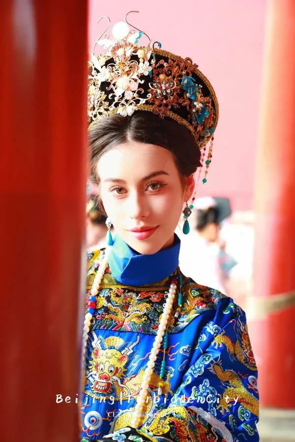 hanfu photoshoot, the forbidden city, beijing, qing dynasty, female, solo, chinatravel, Portrait of a young woman in an ornate gold headdress and blue embroidered traditional robe, framed by red pillars on a pink background.
