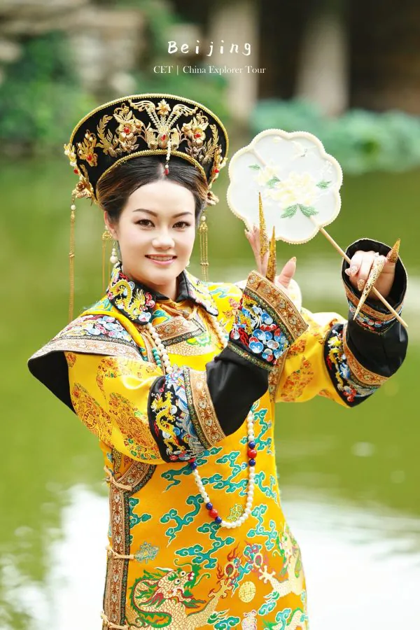 hanfu photoshoot, the forbidden city, beijing, qing dynasty, female, solo, chinatravel, Woman in a bright yellow traditional Chinese imperial costume with a jeweled headdress, holding a decorative fan outdoors by a lake.