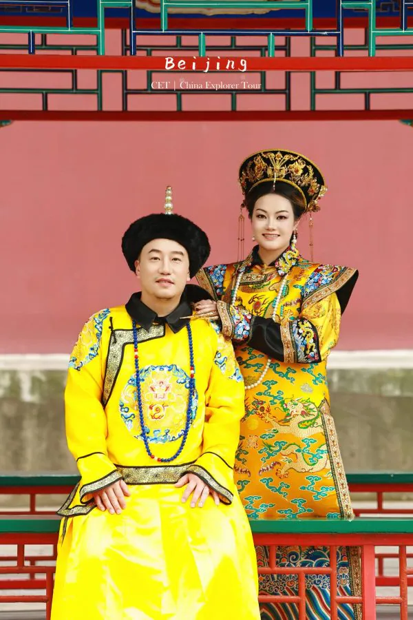 hanfu photoshoot, the forbidden city, beijing, qing dynasty, couple, chinatravel, Couple in ornate yellow Qing dynasty costumes; woman with a gold headdress, man in a black fur hat, posing together in front of a pink wall and decorative lattice railing.