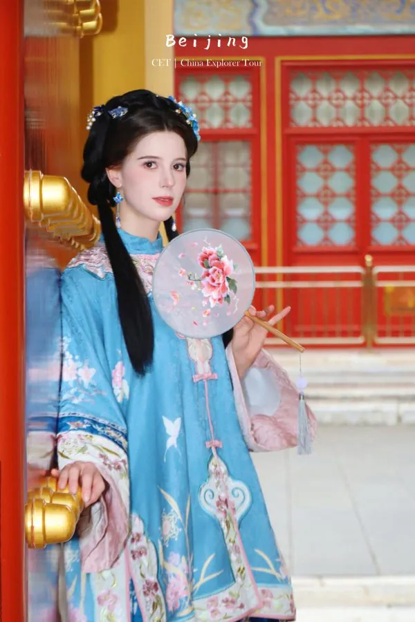 hanfu photoshoot, the forbidden city, beijing, qing dynasty, female, solo, chinatravel, Woman in a blue traditional Chinese dress holding a floral round fan, standing beside a red and gold doorway.