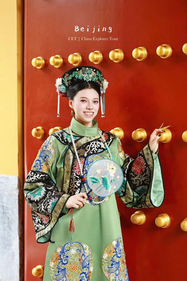 hanfu photoshoot, the forbidden city, beijing, qing dynasty, female, solo, chinatravel, Young woman in an ornate green traditional Chinese dress with a jeweled headdress, standing beside a red door with gold knobs.