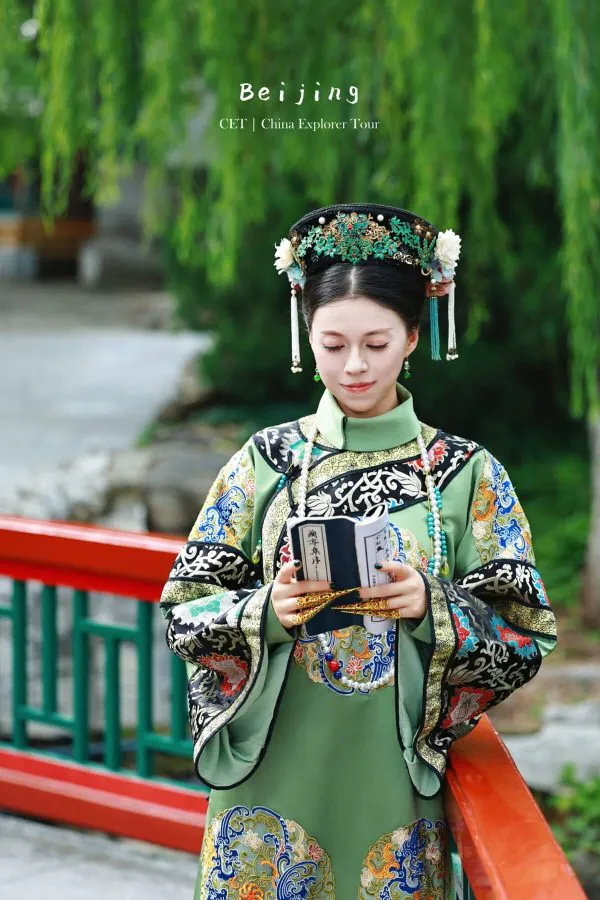 hanfu photoshoot, the forbidden city, beijing, qing dynasty, female, solo, chinatravel, Young woman in an ornate green traditional Chinese outfit and headdress reads a small book outdoors by a red railing and greenery.