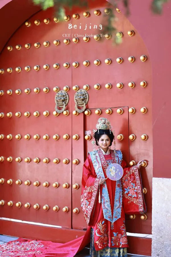 Hanfu photoshoot, zhongshan park, beijing, chinatravel, Woman in a bright red traditional Chinese wedding dress with elaborate headdress stands by a large red door studded with gold knobs, Beijing label visible.