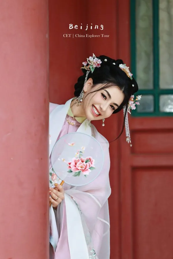 hanfu photoshoot, the forbidden city, beijing, ming dynasty, female, solo, chinatravel, Smiling woman in a pink traditional dress with ornate hair accessories peeks from behind a red pillar, holding a floral fan.