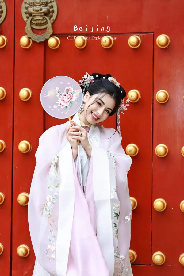 hanfu photoshoot, the forbidden city, beijing, ming dynasty, female, solo, chinatravel, Young woman in pastel pink hanfu holding a floral round fan, smiling in front of a red door with gold knobs.