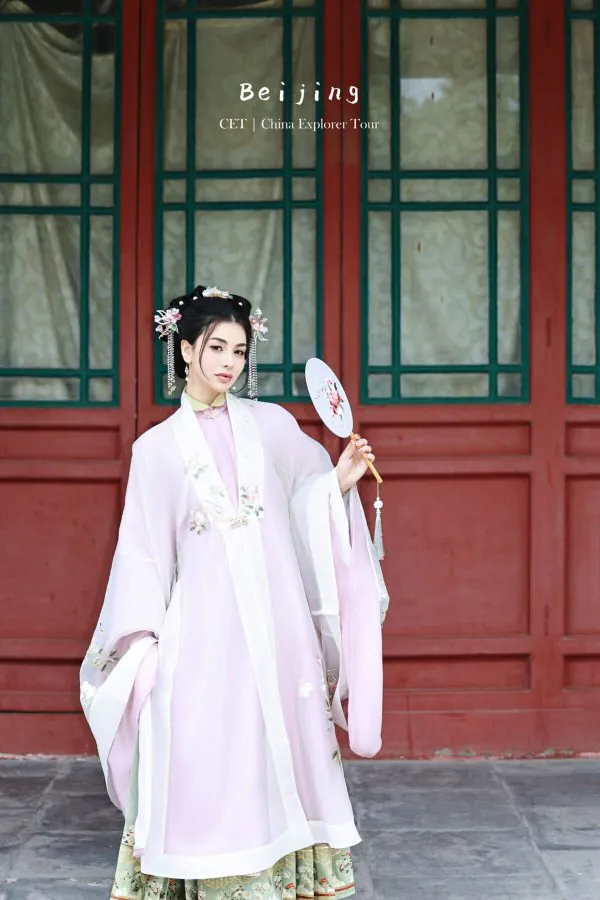 hanfu photoshoot, the forbidden city, beijing, ming dynasty, female, solo, chinatravel, Woman in a pale pink traditional Chinese dress with floral embroidery, standing before a red lattice door and holding a round floral fan.