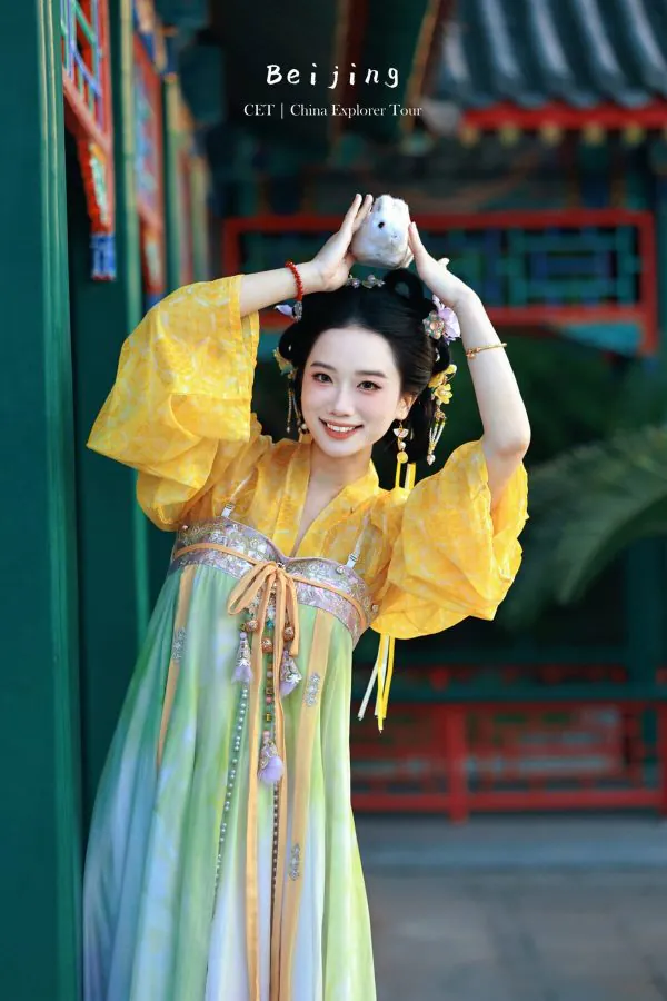 Hanfu photoshoot, the forbidden city, beijing, chinatravel, Smiling woman in a yellow traditional dress posing with hands above her head, holding a white fluffy accessory, in a temple courtyard.