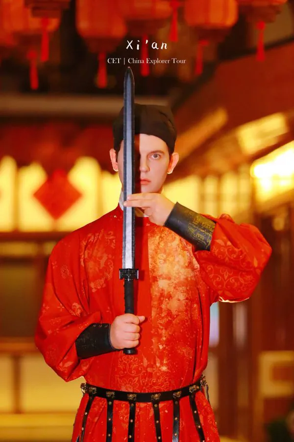 Hanfu photoshoot, xingqing palace, xi'an, solo, male, chinatravel, Performer in red ceremonial robes holds a vertical sword up to their face in a staged pose, obscuring half of their face.