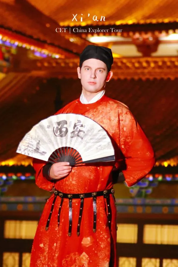 Hanfu photoshoot, xi'an, xingqing palace, male, chinatravel, Man in a red traditional Chinese outfit wearing a black hat, holding a white fan with calligraphy in front of a lit temple at night.