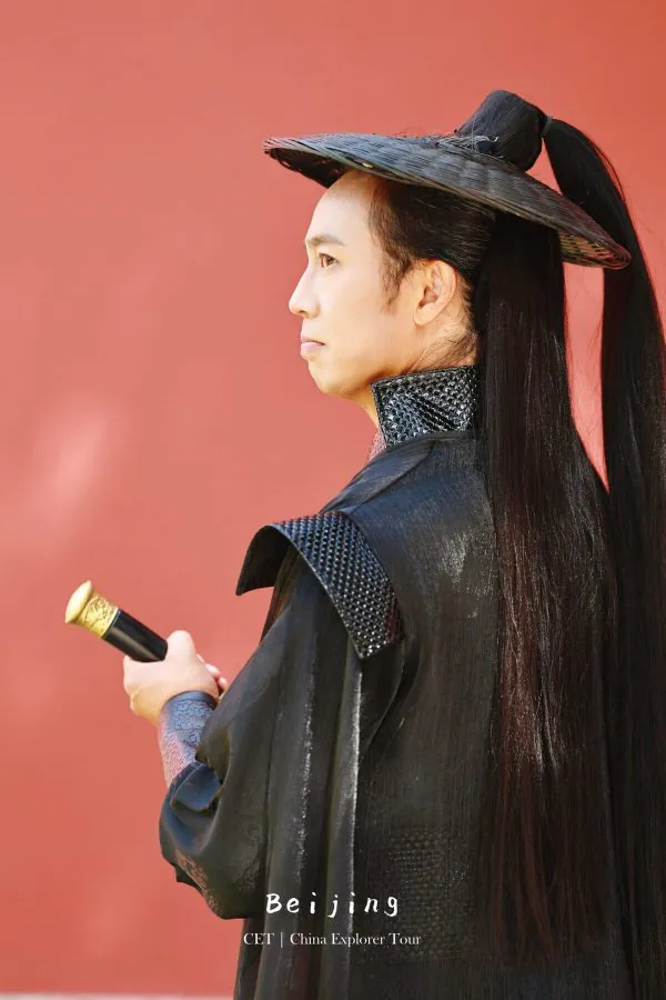 Hanfu photoshoot, the forbidden city, beijing, chinatravel, Person in traditional Chinese costume with a wide-brimmed hat, long black hair, and armor-like clothing against a red wall, holding a ceremonial baton.