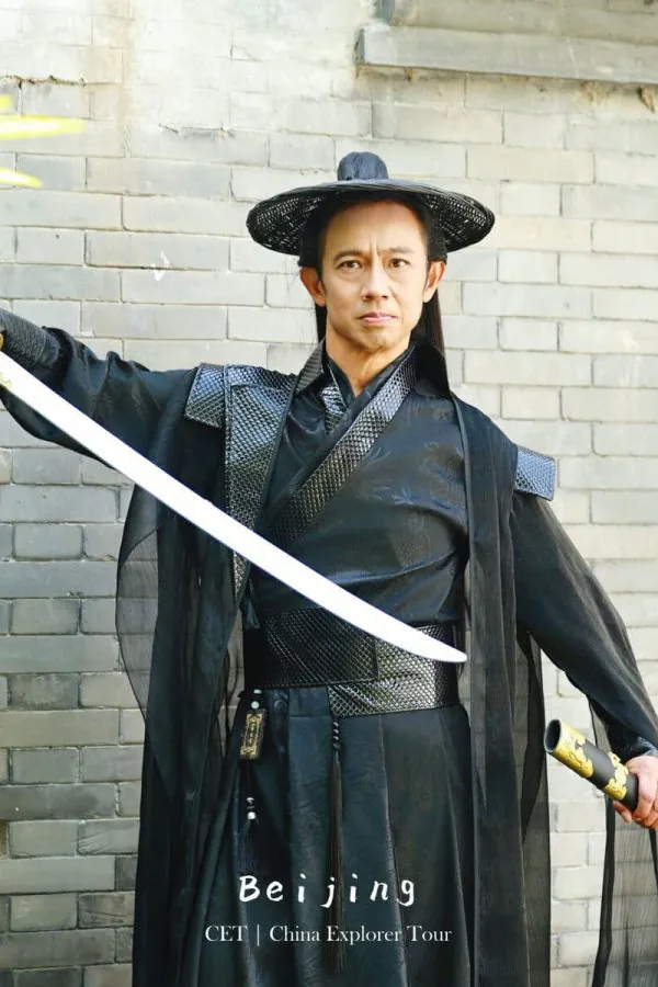 Hanfu photoshoot, the forbidden city, beijing, chinatravel, Man in a black traditional Chinese robe holding a sword, posing against a gray brick wall; Beijing travel promo text visible at bottom.
