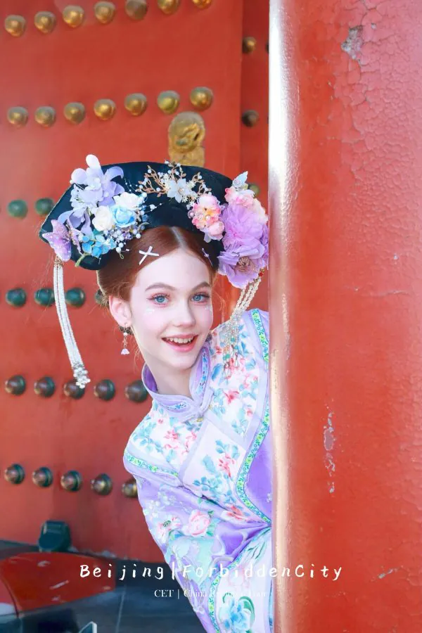 hanfu photoshoot, the forbidden city, beijing, qing dynasty, female, solo, chinatravel, Young woman in a purple floral qipao with an elaborate flower headdress peeks from behind a red pillar at Beijing's Forbidden City, with 'Beijing Forbidden City' text visible.