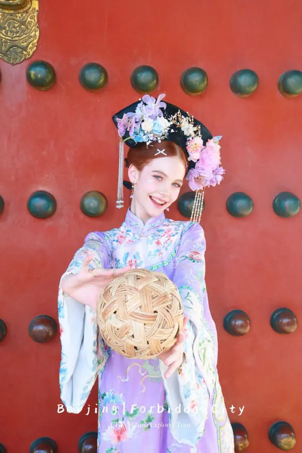 hanfu photoshoot, the forbidden city, beijing, qing dynasty, female, solo, chinatravel, Young woman in a pastel floral hanfu with an ornate flower headdress, extending a woven ball toward the camera against a red gate backdrop.