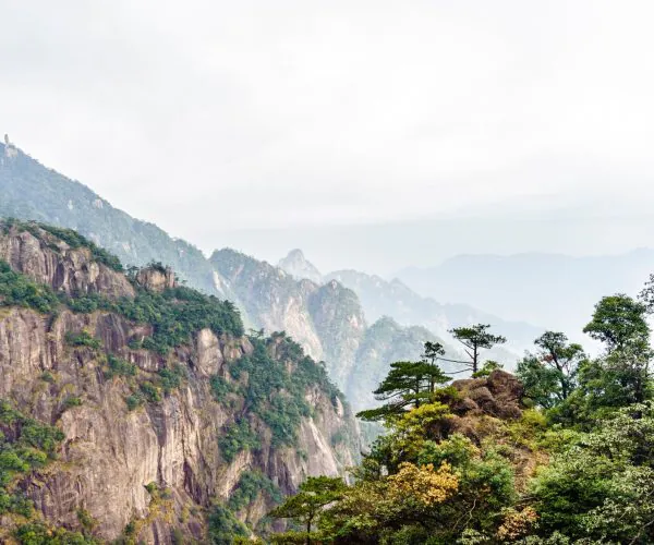 Shangrao Weather,Shangrao,Chinatrip Rocky mountain cliffs covered with trees, fading into a hazy blue horizon under a pale sky.