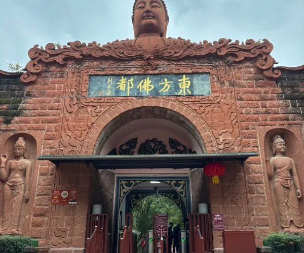 Oriental Buddha Park,Leshan,Chinatravel Temple entrance with a large Buddha head statue atop a red, intricately carved archway and gold Chinese characters above the doorway, flanked by stone statues; visitors enter through an open gate.