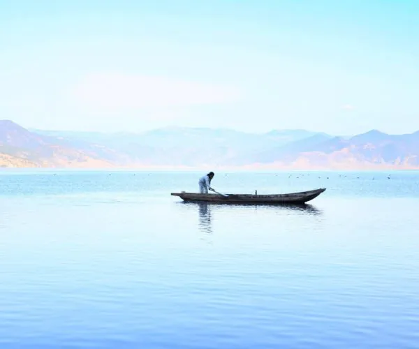 Lijiang Weather,Lijiang,Chinatravel Person in a small wooden boat paddling on a calm lake with distant mountains in the background.