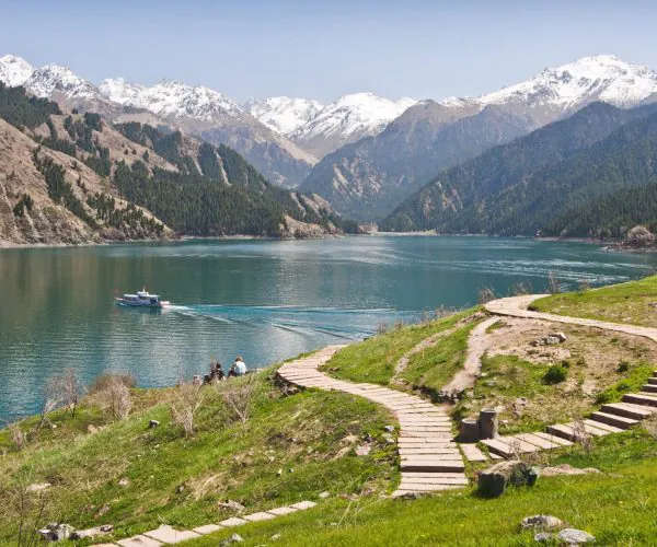 Urumqi,Xinjiang,Chinatrip Turquoise lake with snow-capped mountains, a small boat gliding on the water, and a stone path winding along a grassy hillside where a few people sit nearby.