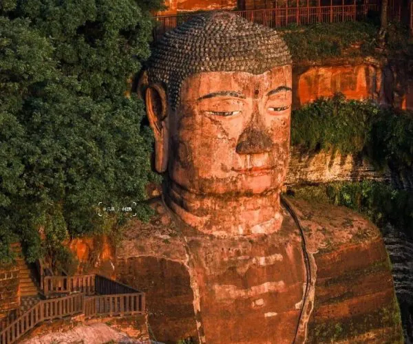 Leshan Giant Buddha,Leshan,Chinatravel Giant stone Buddha head carved into a red rock cliff, surrounded by dense green trees and a wooden walkway nearby