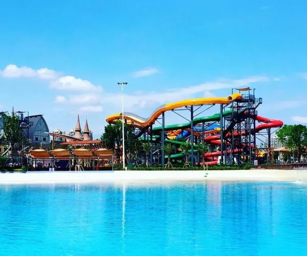 Leshan Happy Water World,Leshan,Chinatravel Colorful winding water slides at a sunny outdoor water park beside a large blue pool in front of palm trees and buildings.