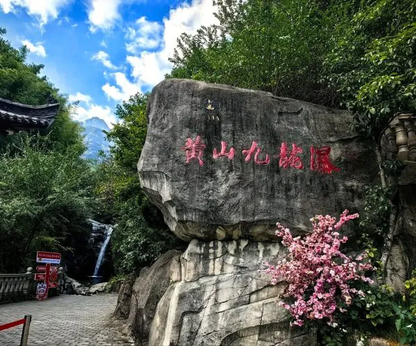 Nine Dragon Waterfall,Huangshan,Chinatravel Large rock with red Chinese characters beside a flowering pink bush in a lush garden with a small waterfall and paved path.