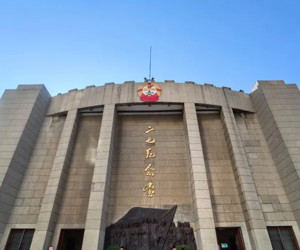 Erqi Memorial Tower,Zhengzhou,Chinatravel Large gray stone building with tall arches and golden vertical Chinese characters on the façade, a crest atop the center, and a bas-relief sculpture near the entrance against a clear blue sky