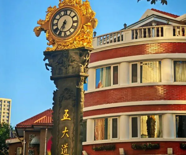 Ornate gold clock on a carved dark pillar with Chinese characters, beside a curved red-brick building under a blue sky.