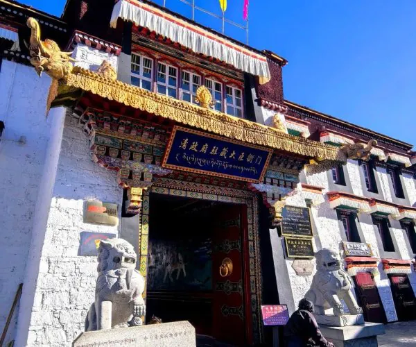 Barkhor Street Old Town,Lhasa,Chinatravel Tibetan temple entrance with ornate gold decorations, colorful flags, and stone guardian lions as a monk with a walking stick stands nearby against a clear blue sky.