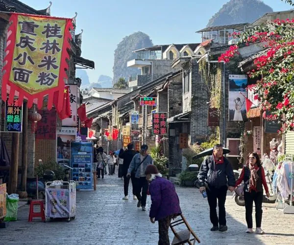 Xingping Ancient Town,Yangshuo,Chinatravel Narrow historic street in a Chinese town with stone pavement, red banners, and shoppers; mountains in the background.