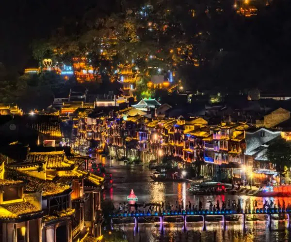 Fenghuang Ancient Town,Fenghuang,Chinatravel Nighttime riverside town with lantern-lit buildings, a crowded promenade, and reflections on the water.