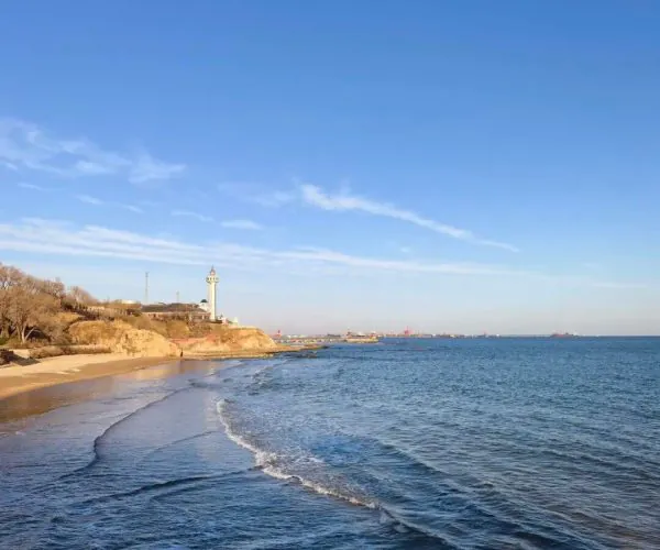 Beidaihe Seaside Resort,Qinhuangdao,Chinatravel Coastal scene with a sandy beach, rocky shoreline, and a lighthouse on the cliff to the left under a clear blue sky