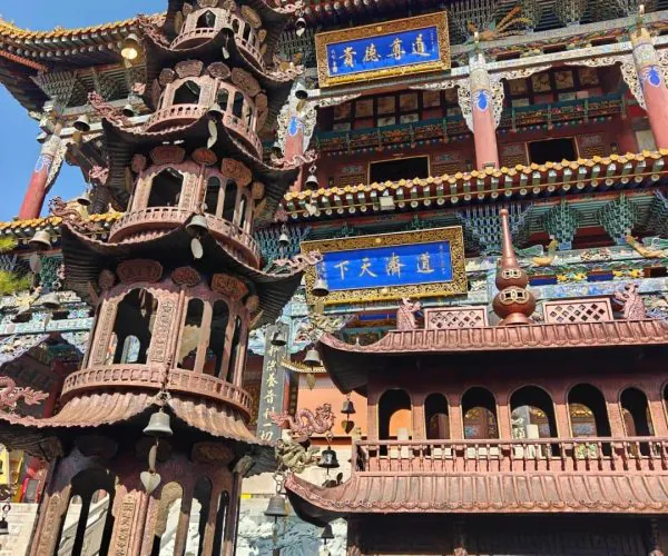 Tulou Temple Tibetan Buddhist Sites,Xining,Chinatrip Intricately carved multi-tier pagoda in front of a colorful traditional Chinese temple with blue plaques and ornate roofs under a clear sky.