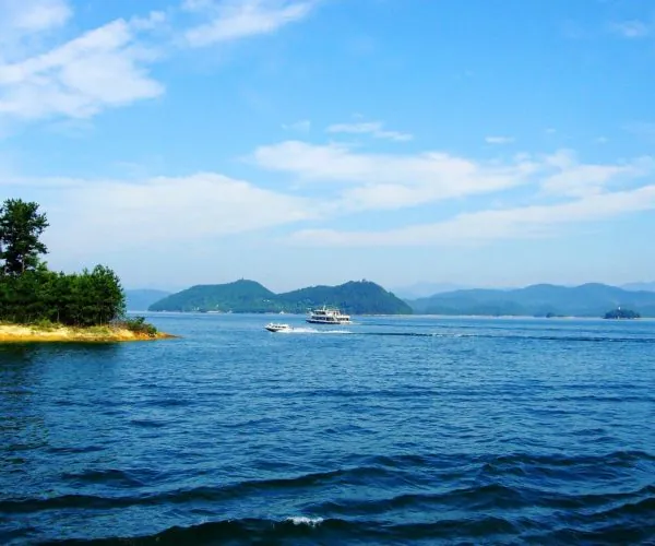 Thousand Islands Lake,Hangzhou,Chinatravel Calm blue sea with a sandy, tree-lined shore on the left and two boats crossing toward distant island hills far ahead.