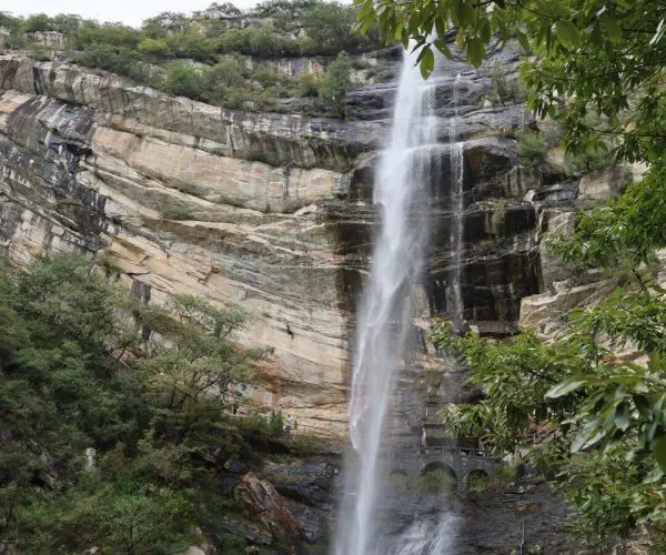 Luya Waterfall,Shaolin & Songshan Area,Chinatrip Tall waterfall cascading down a layered rocky cliff into a pool, surrounded by green trees and boulders steadily misting the area.