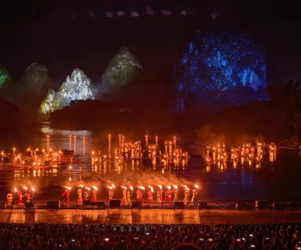 Impression Sanjie Liu Show,Yangshuo,Chinatravel Torch-lit water performance at night: performers with flames on a lake, audience in foreground, with colored rock formations lit in green, yellow, and blue.