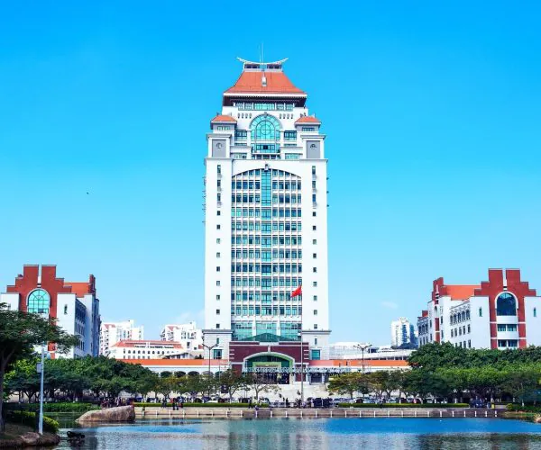 Xiamen University,Xiamen,Chinatravel Tall white campus tower with a red-tiled roof, flanked by smaller red-roofed buildings under a clear blue sky, with trees and a pond in front.