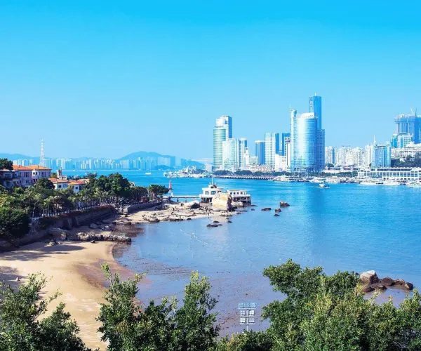 Xiamen Weather,Xiamen,Chinatravel Coastal city skyline across a calm bay with modern high-rise buildings, a sandy beach in the foreground, and green trees along the shore.