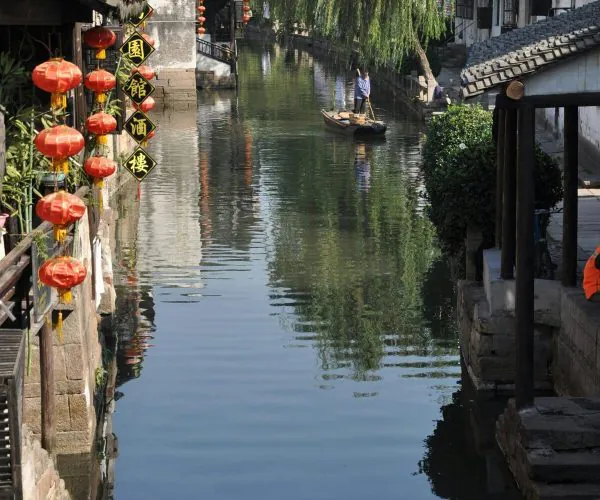Nanchan Temple and Ancient Town,Suzhou,Chinatravel Canal flanked by traditional white-walled buildings with red lanterns, a man paddling a small boat under a weeping willow.