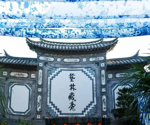 Zhoucheng Village Bai Culture Museum,Dali,Chinatravel Traditional Chinese gateway with ornate grey stone panels, blue-white calligraphy panels, and a wooden wine barrel on a stepped platform beneath blue tie-dye banners.