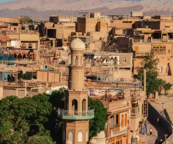 Kashgar Old Town,Kashgar,China tour Cityscape with beige, adobe-style buildings and a tall ornate tower with a domed top in the foreground; desert mountains in the distance under a clear blue sky.