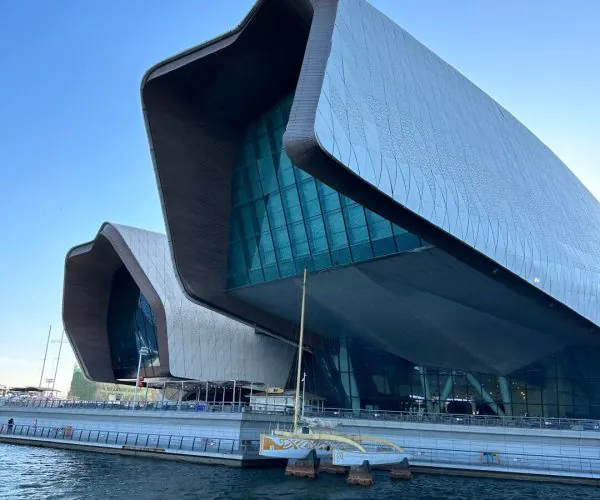 Modern waterfront concert hall with a sweeping curved silver roof and glass facade by calm blue water.