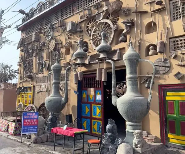 Karez Folklore Garden,Turpan,China tour Eclectic storefront wall filled with metal sculptures and vintage tools, with colorful doors and a street-side table and chair up front.