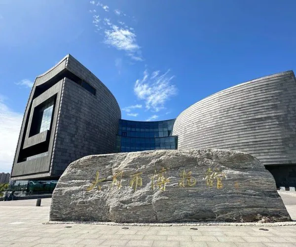 Datong Museum,Datong,Chinatravel Exterior of Dalian City Museum: a modern, curved gray stone building with a large rock in the foreground inscribed in gold Chinese characters under a blue sky.