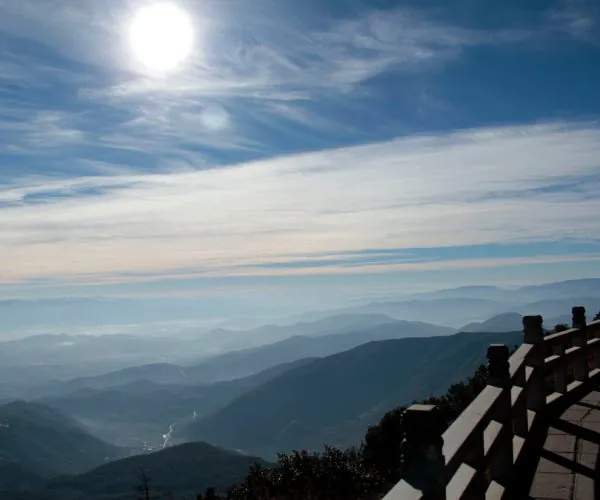 Dali Weather,Dali,Chinatravel Mountain range under a bright sun with wispy clouds and a wooden railing in the foreground.