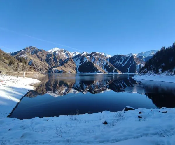 Heavenly Lake of Tianshan,Urumqi,Chinatrip nowy shoreline and a calm lake reflecting snow‑capped mountains under a clear blue sky.